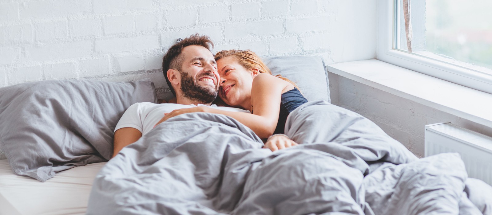 Paar liegt lächelnd im Bett, umarmt sich unter einer grauen Decke, mit Fenster im Hintergrund.