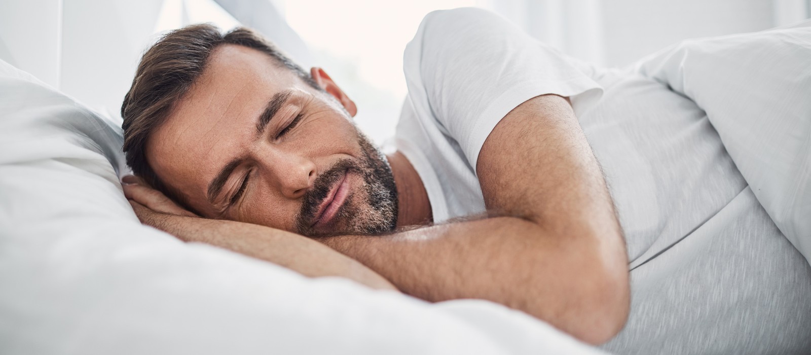 Uomo dorme rilassato su un letto circondato da cuscini e coperte bianche.