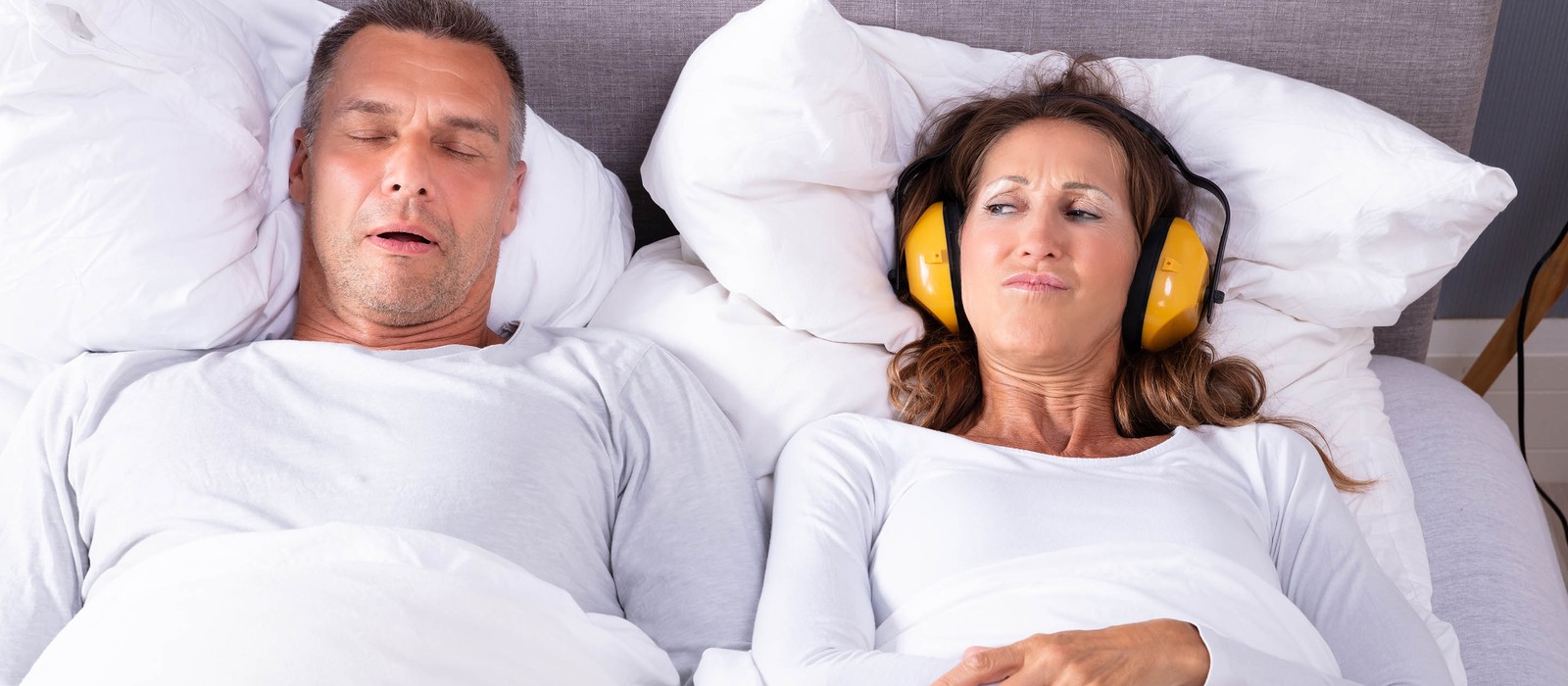 Mann schläft im Bett, während Frau mit Kopfhörern unzufrieden daneben liegt.
