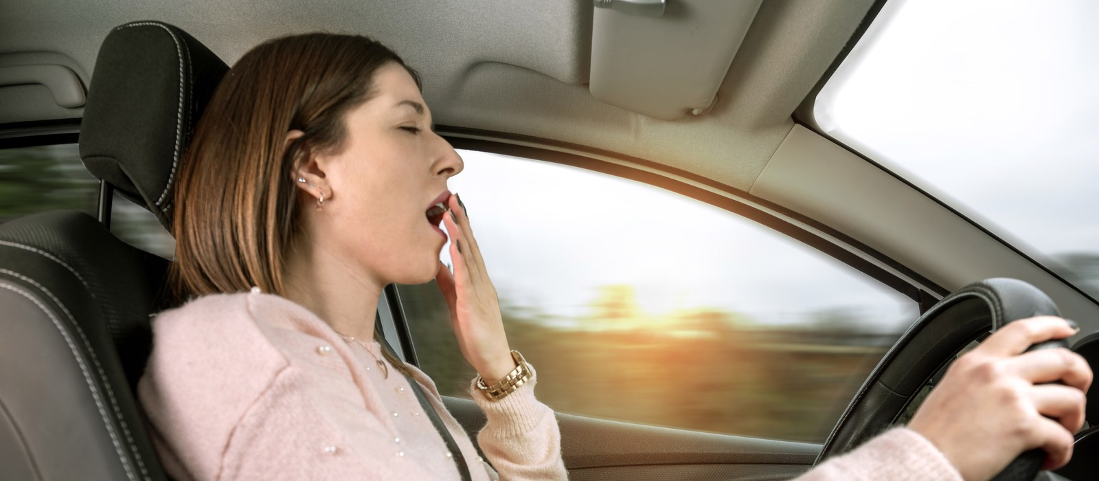 Donna in auto che sbadiglia stanca durante il viaggio, con sfondo sfocato e luce solare.