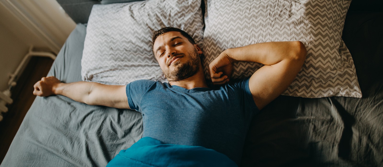 Uomo sdraiato rilassato a letto, circondato da cuscini e una coperta, con un'espressione soddisfatta.