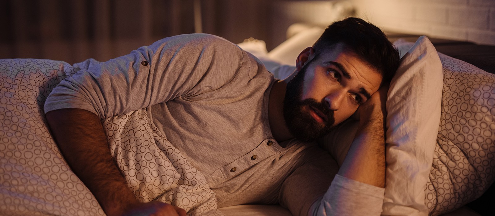 Uomo sdraiato a letto, guarda pensieroso il soffitto, circondato da cuscini e una coperta.