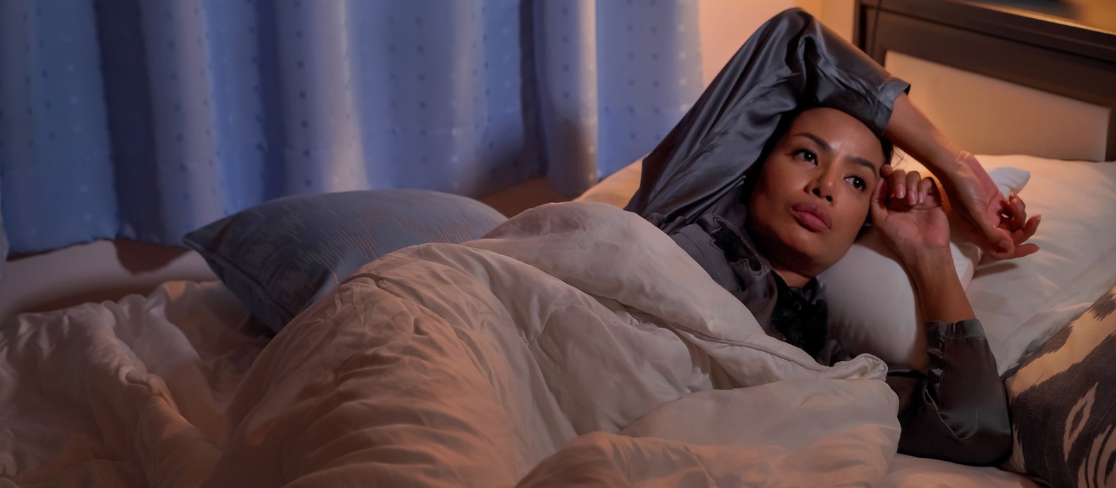 Eine nachdenkliche Person liegt in einem Bett, umgeben von Kissen und einer Decke, mit Vorhängen im Hintergrund.