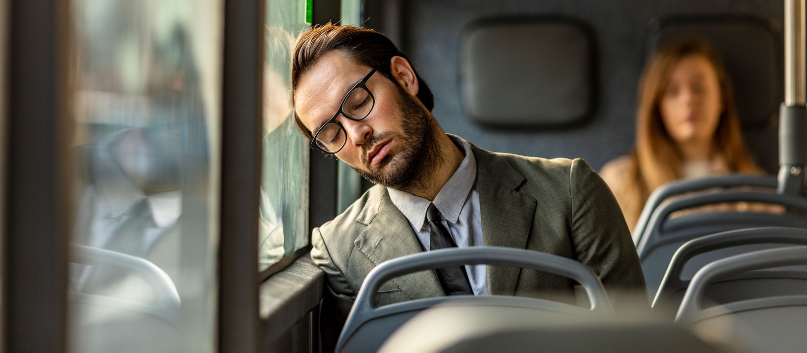 Mann im Anzug schläft im Bus, während eine Frau im Hintergrund sitzt.