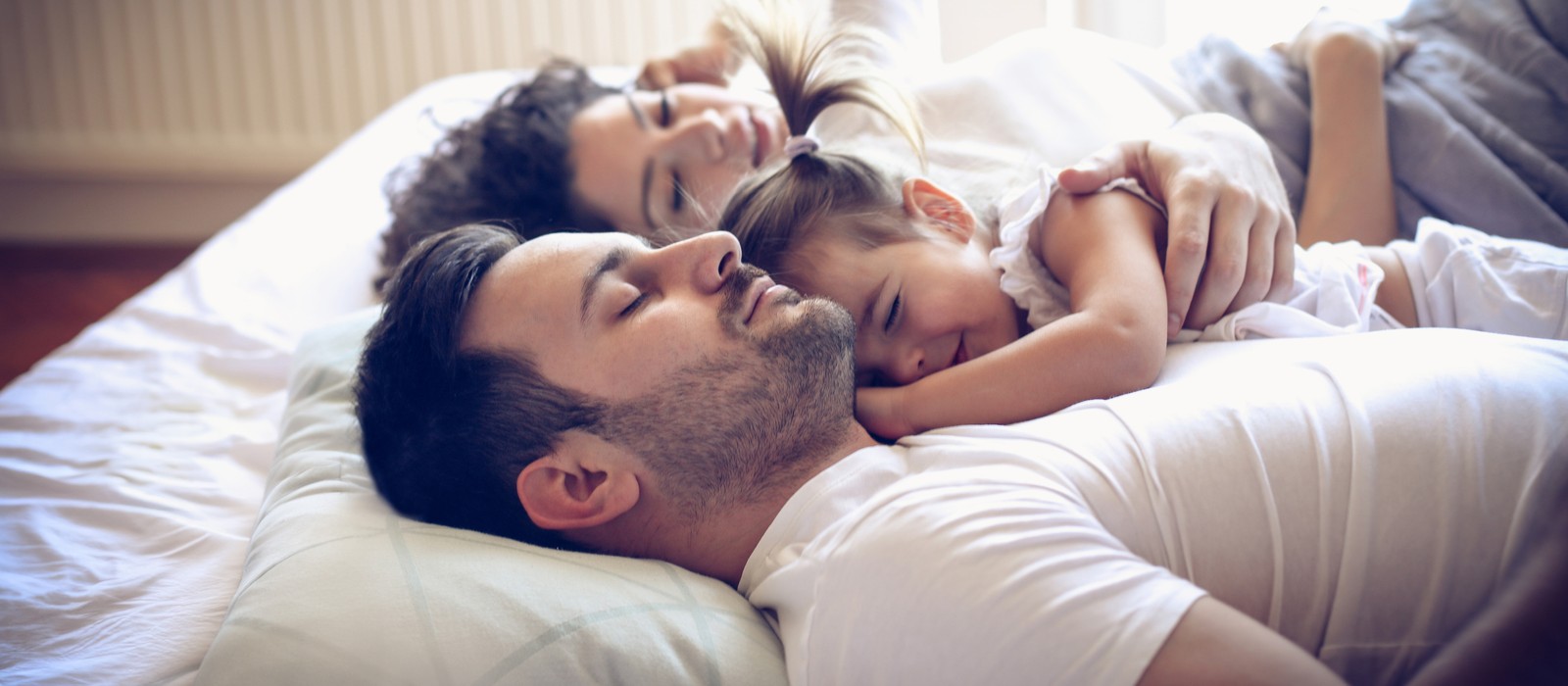 Una famiglia dorme rilassata nel letto, un genitore ha un bambino sul petto.