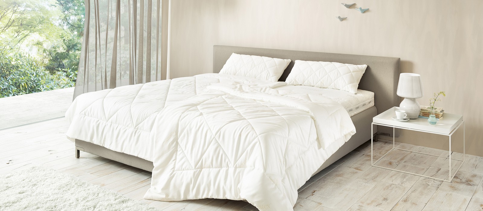 Camera da letto luminosa con letto, copriletto bianco, cuscini e comodino con lampada e vetro.