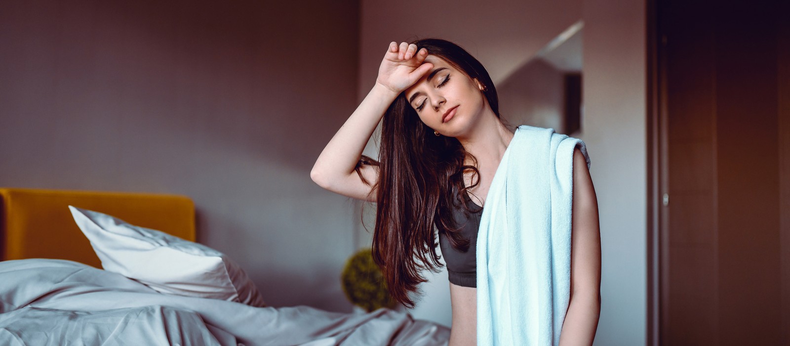 Giovane donna con mano sulla fronte, sembra stanca, in una camera da letto con letto sfatto.