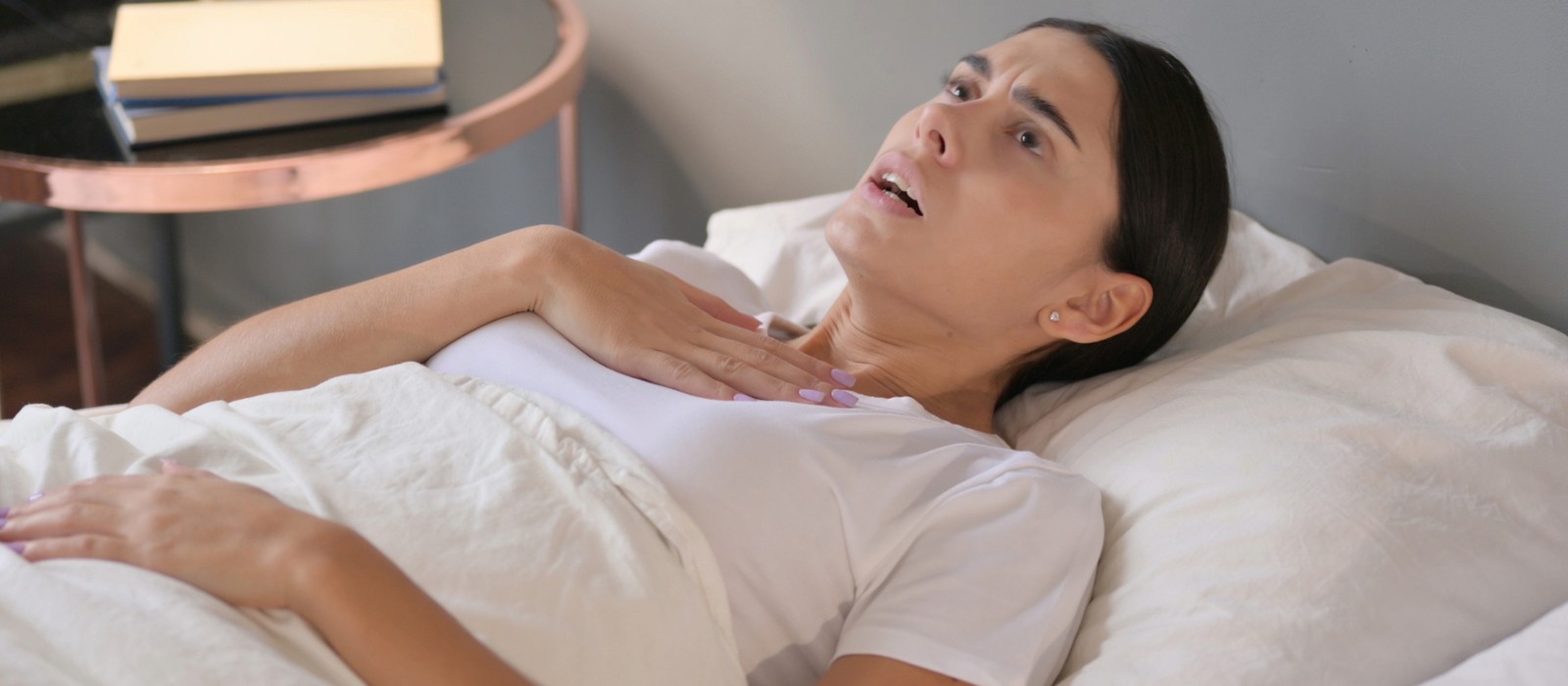 Donna sdraiata a letto, appare spaventata e si tiene la mano sul petto.