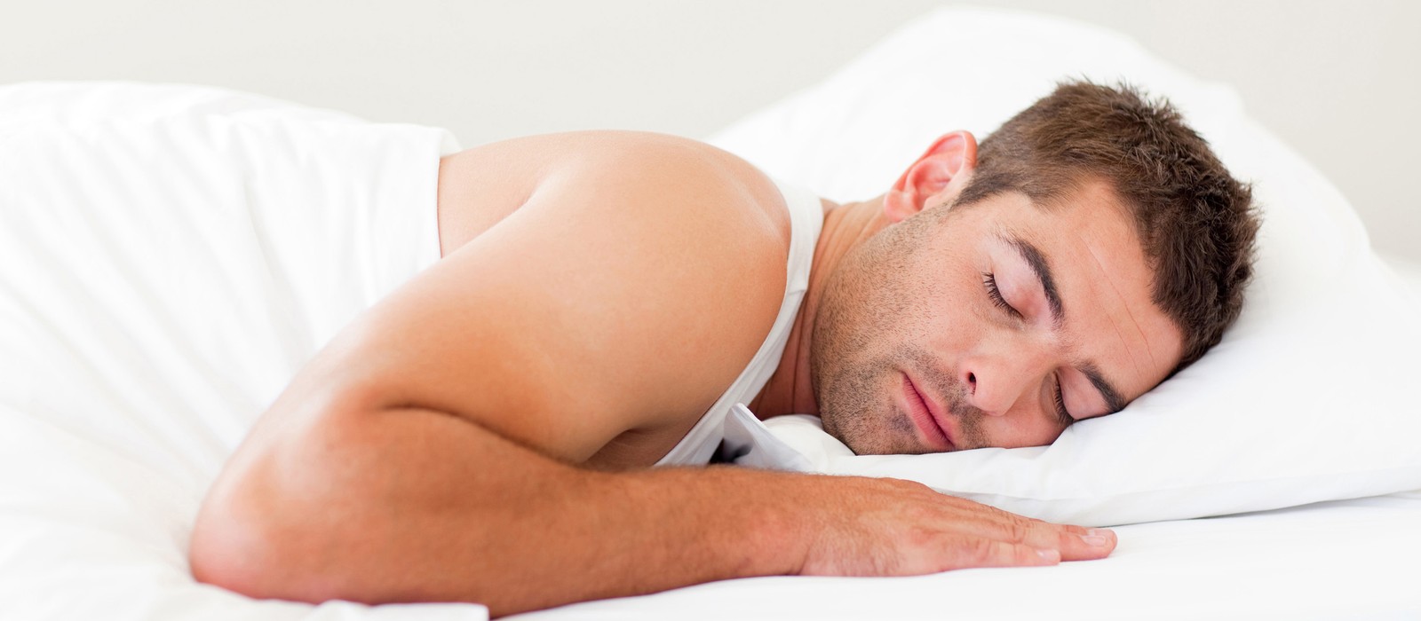 Uomo dorme rilassato su un letto con cuscini e coperte bianche.