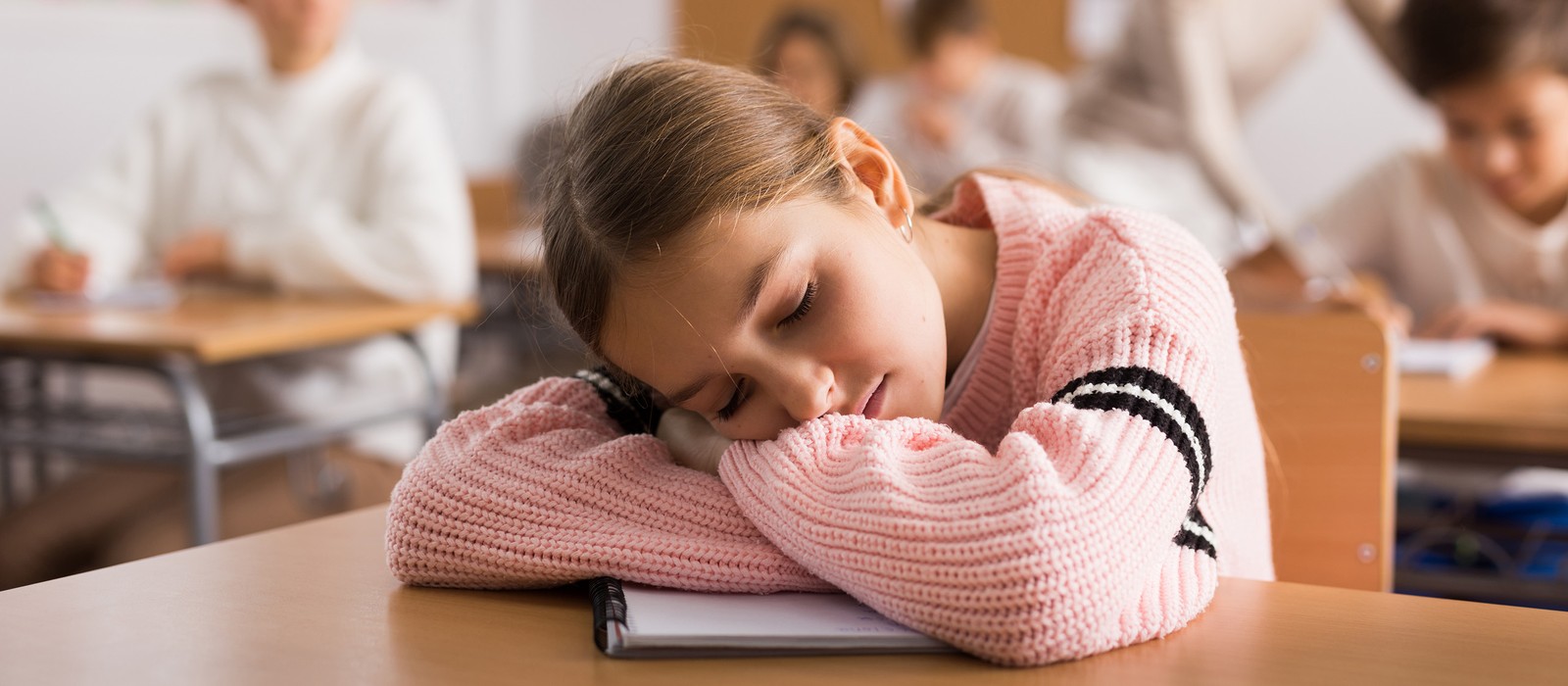 Mädchen mit geschlossenen Augen schläft auf einem Tisch in einem Klassenzimmer. Im Hintergrund sind andere Kinder zu sehen.