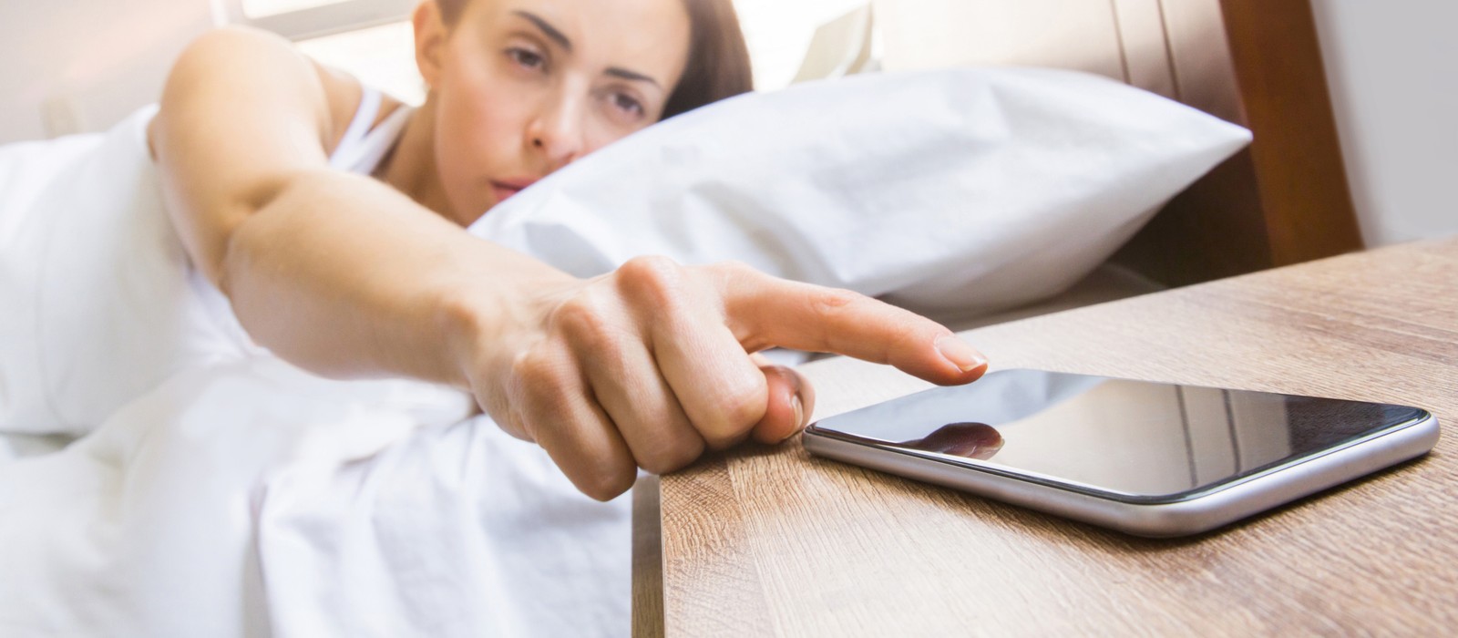 Donna nel letto preme sul display di uno smartphone su un comodino.