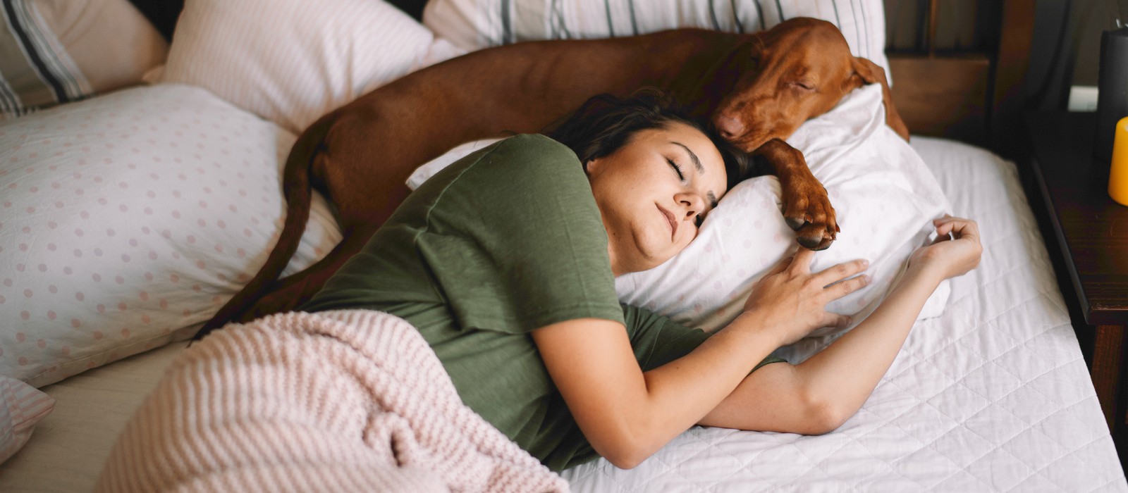 Donna che dorme rilassata nel letto, con un cane sul cuscino, circondata da morbidi biancheria da letto.