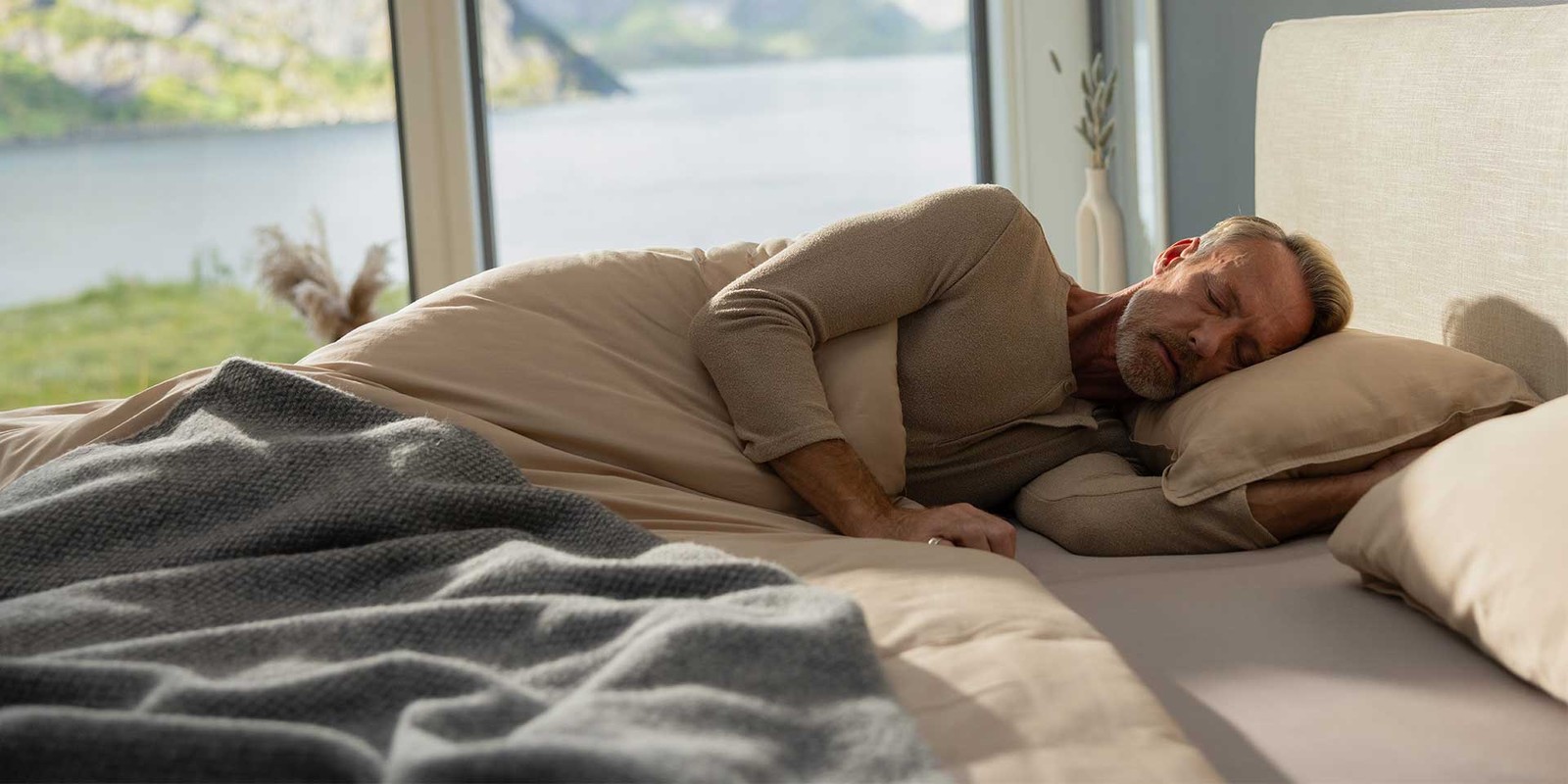 Un uomo anziano dorme rilassato su un letto con cuscini, circondato da un paesaggio tranquillo.