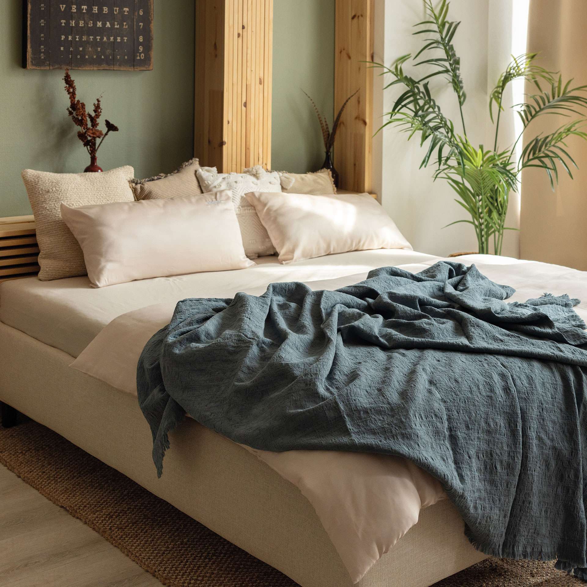 A cozy bedroom with a bed, pillows, a blanket, and plants in the background.
