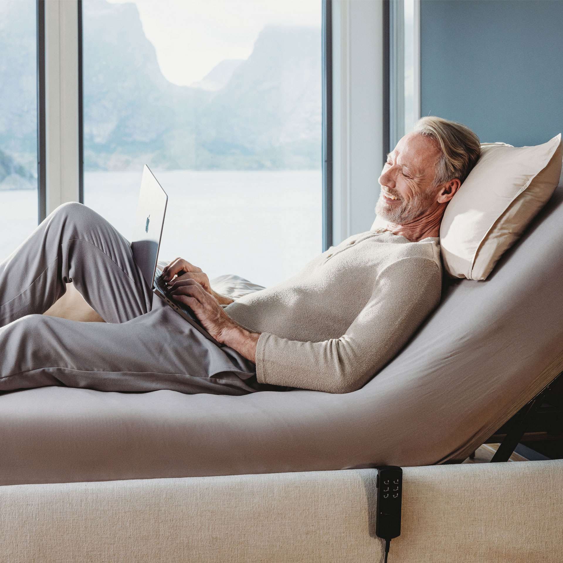 Man relaxing on an adjustable bed, working on a laptop with a view of a landscape.