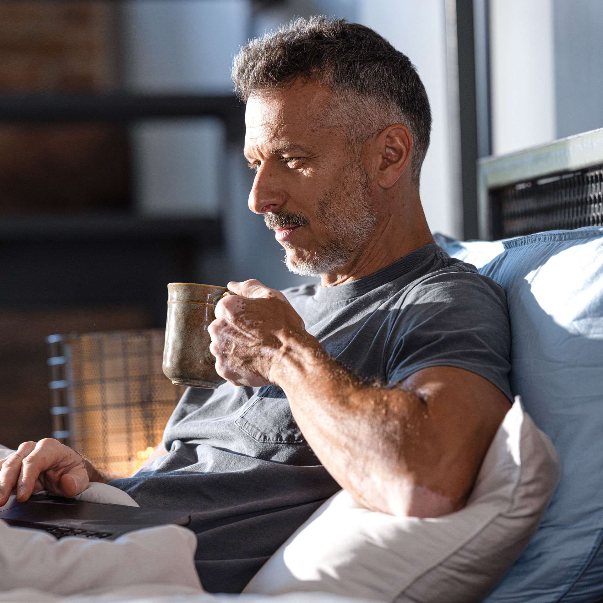 Man sitting on the bed, drinking from a cup and looking at a laptop. Cozy atmosphere with daylight.