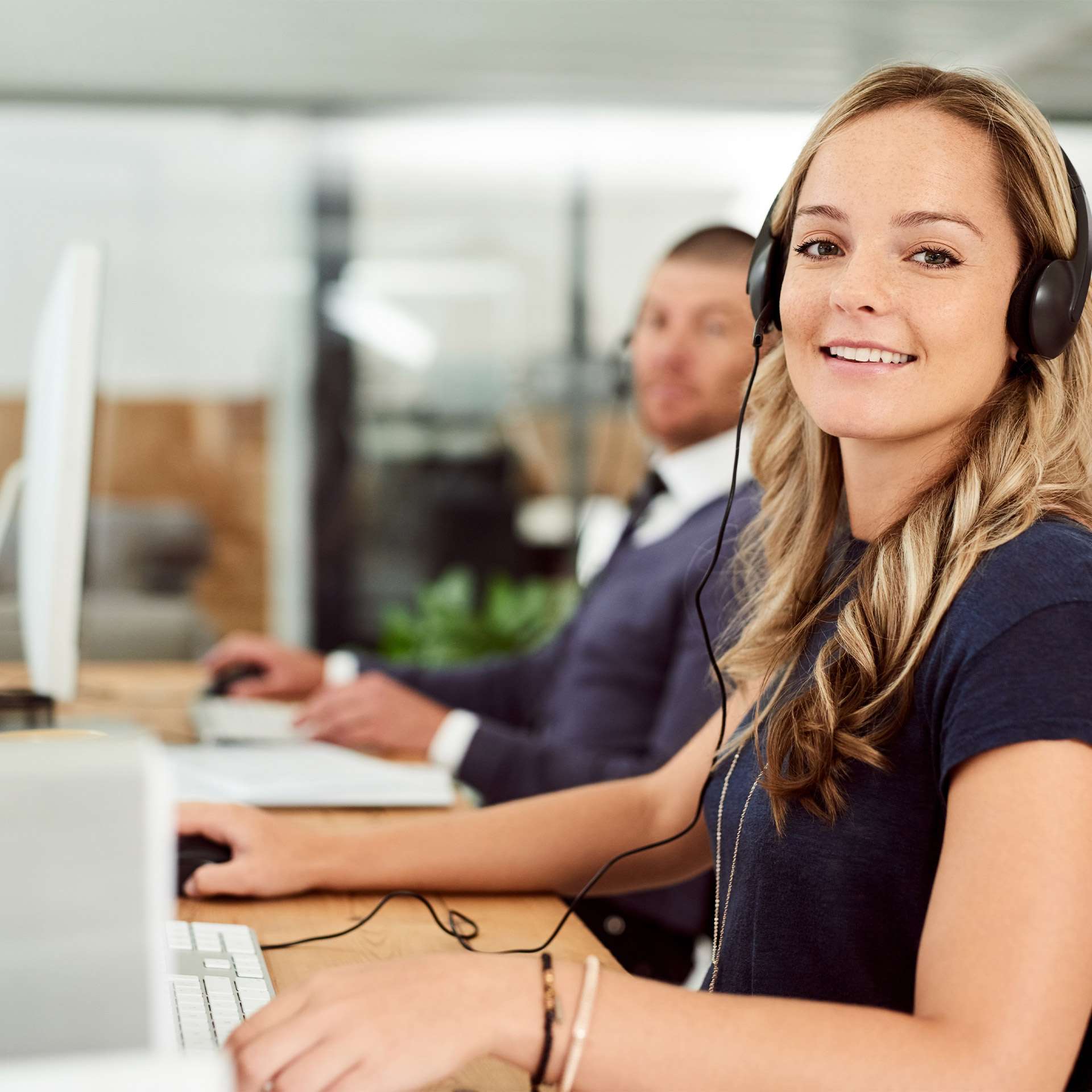 Frau mit Headset am Schreibtisch, im Hintergrund ein Mann an einem Computer. Moderne Büroatmosphäre.