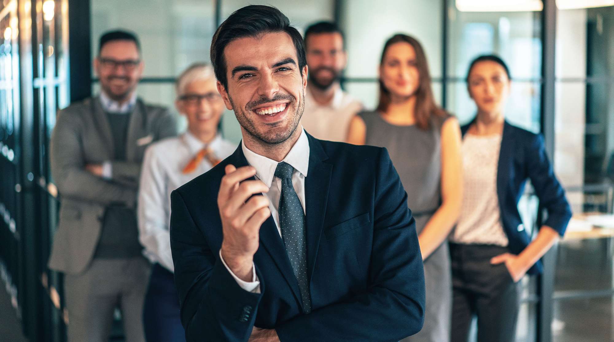Uomo sorridente in giacca davanti a un gruppo di persone in un ufficio moderno.