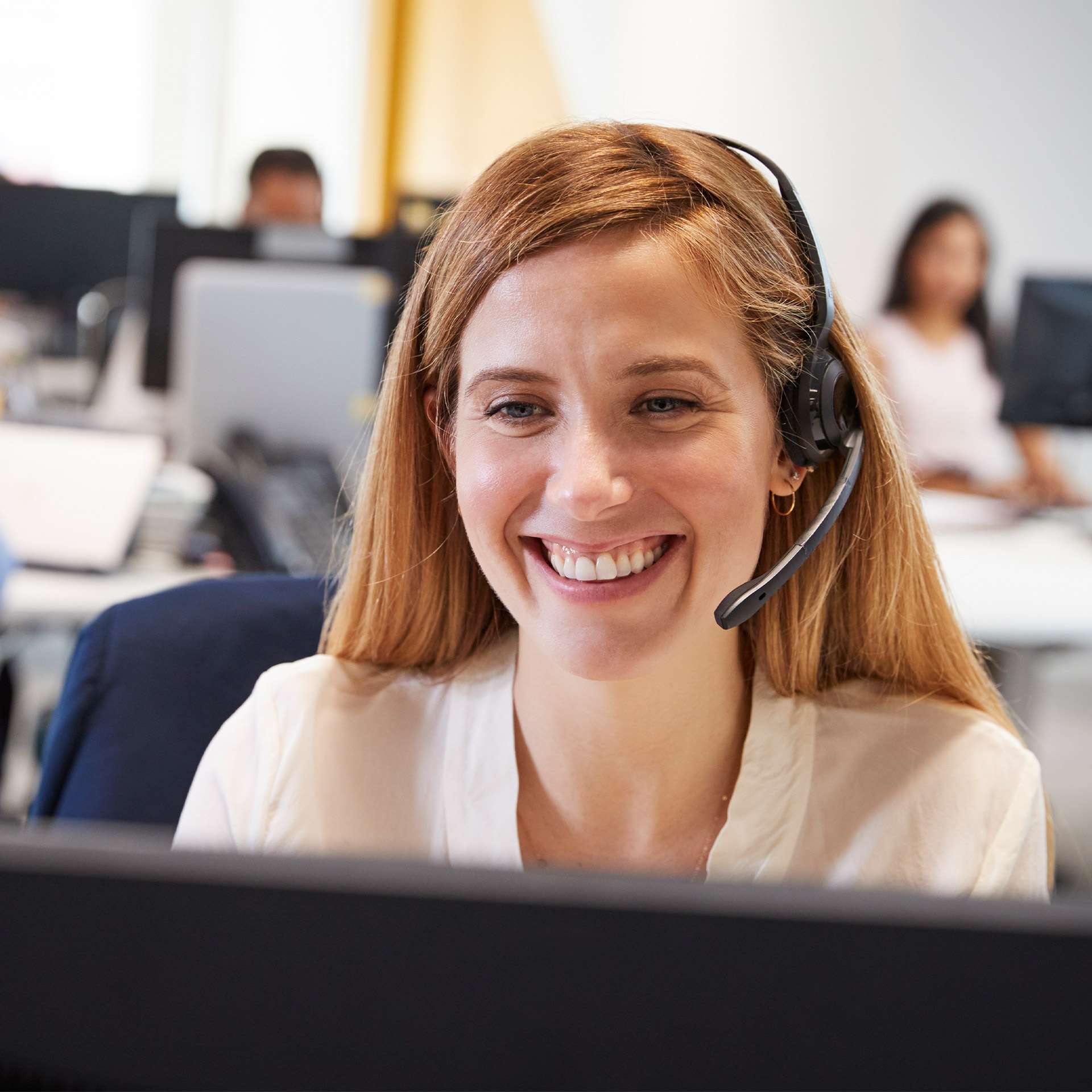 Frau mit Headset lächelt am Schreibtisch in einem modernen Büro, im Hintergrund weitere Mitarbeiter.