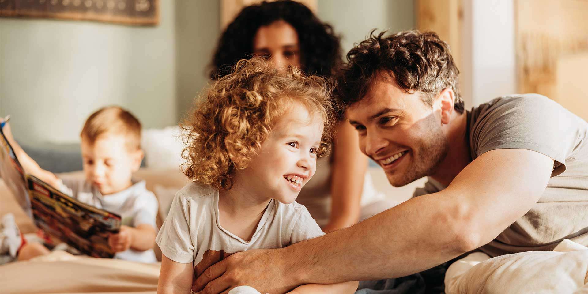 Familie mit zwei Kindern auf einem Bett, lächelnd und spielend in einem gemütlichen Schlafzimmer.