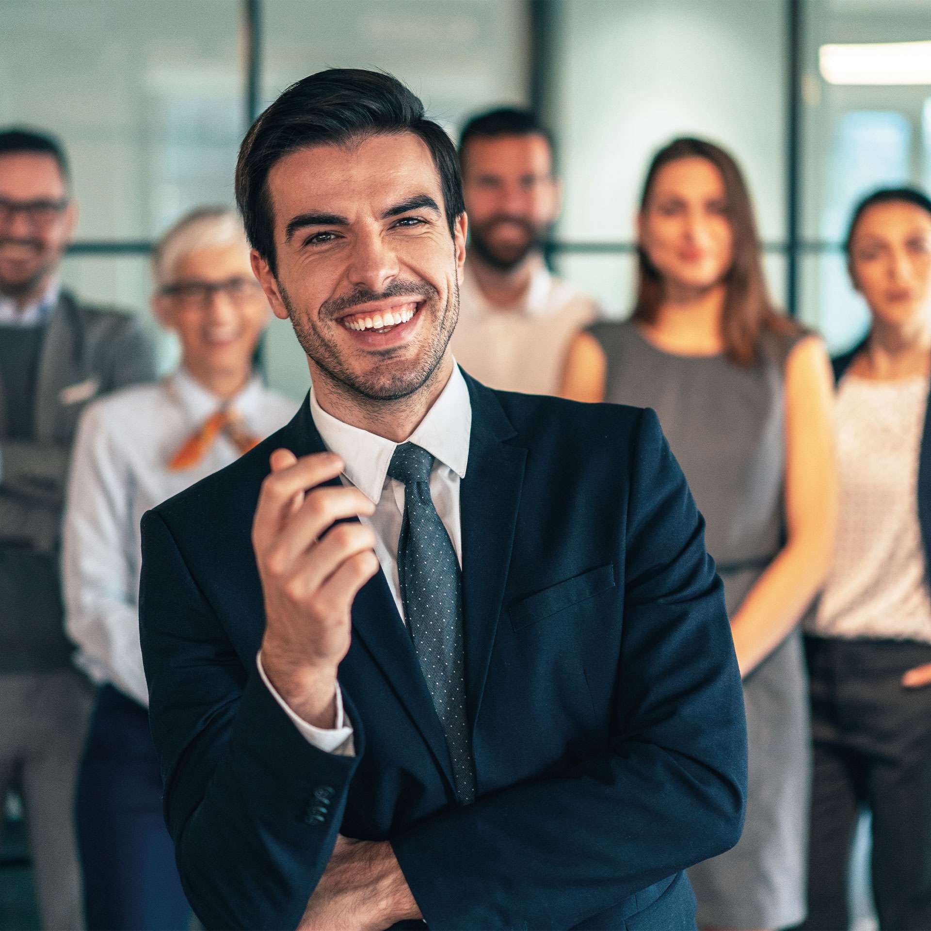 Uomo sorridente in giacca e cravatta davanti a un gruppo di cinque persone in un moderno ufficio.