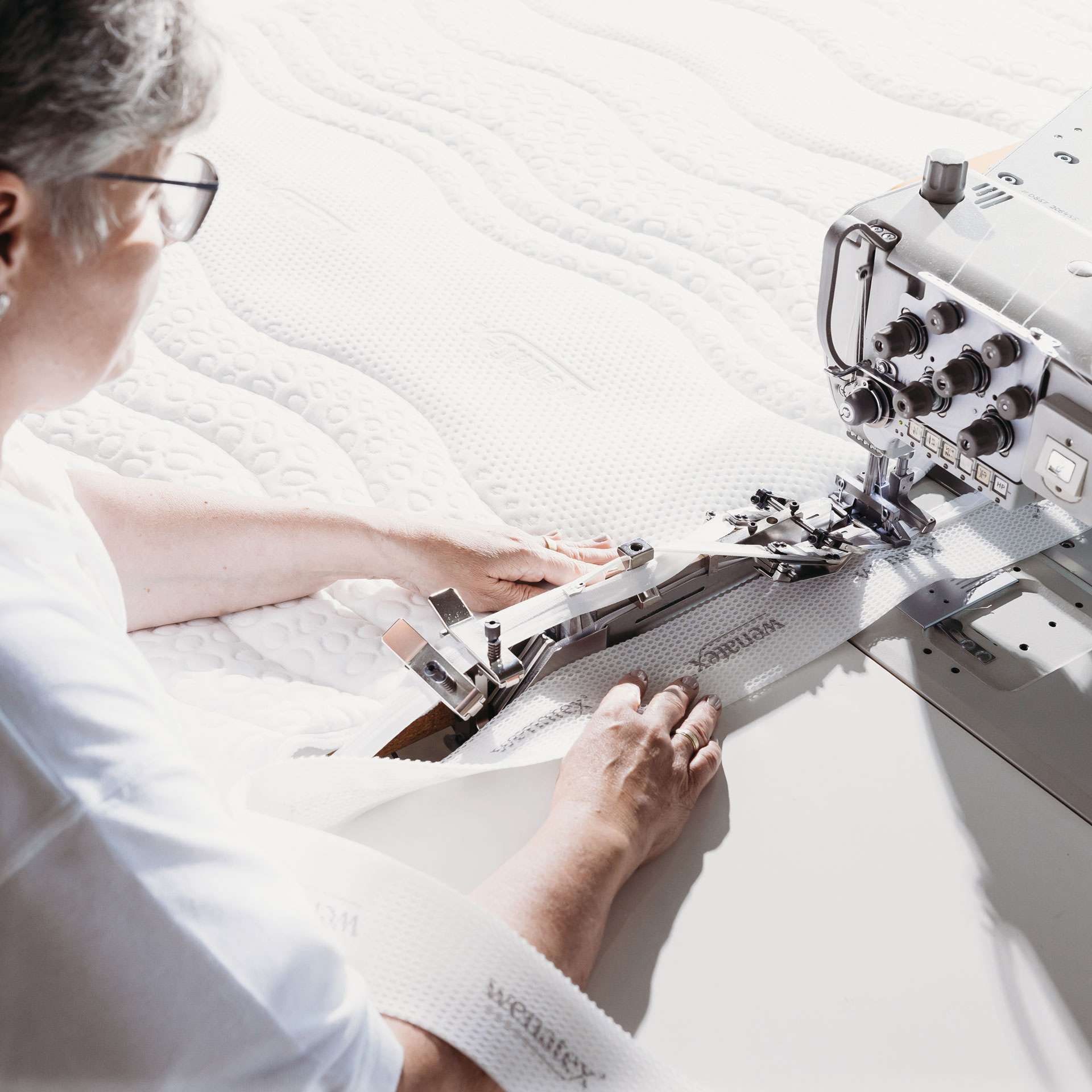 Frau näht einen Matratzenbezug an einer industriellen Nähmaschine. Im Hintergrund ist eine Matratze sichtbar.