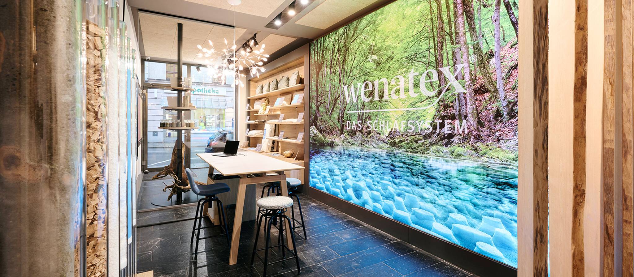 Innenansicht eines modernen Raumes mit Holzregalen, Tisch und großem Wandbild einer Waldlandschaft.