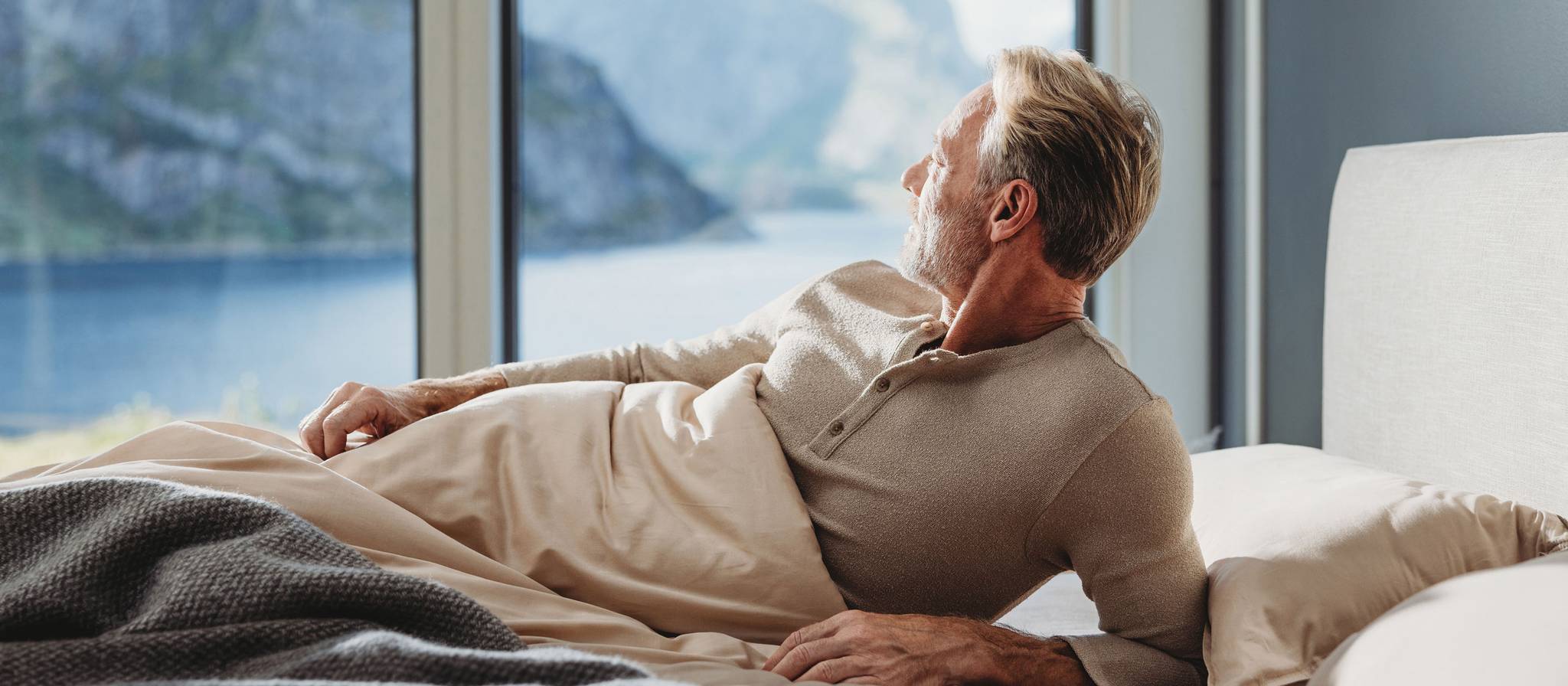 Uomo sdraiato nel letto che guarda fuori dalla finestra su un paesaggio pittoresco.