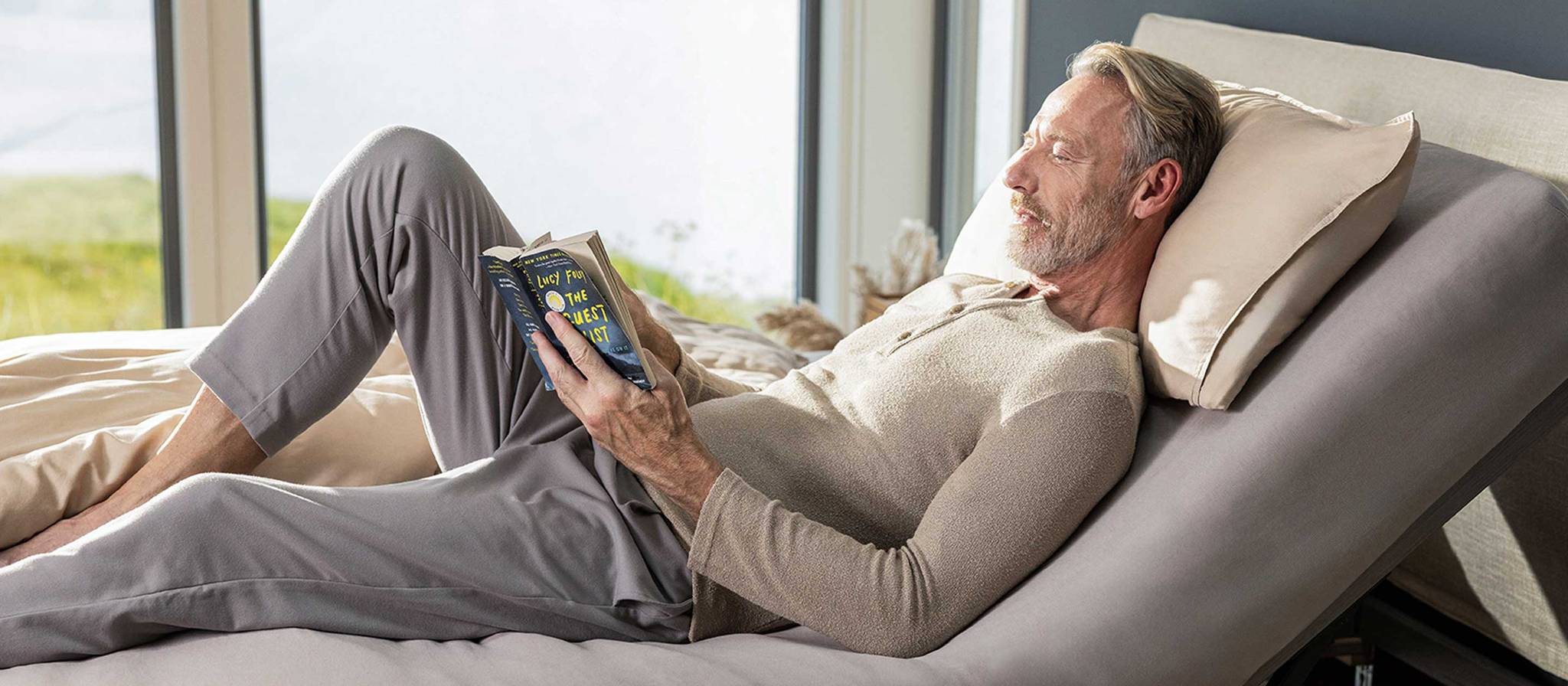 Mann liest entspannt im Bett, umgeben von einer gemütlichen Bettdecke und Kissen.