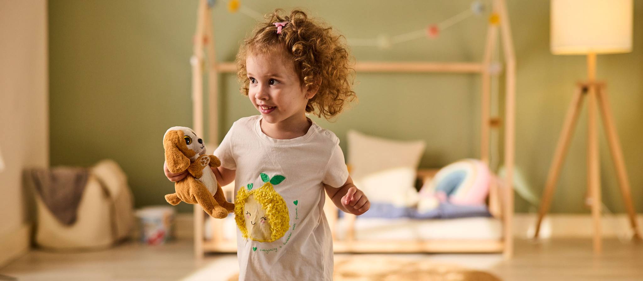 Lächelndes Kind mit Spielzeug in einem gemütlichen Kinderzimmer mit einem Bett im Hintergrund.