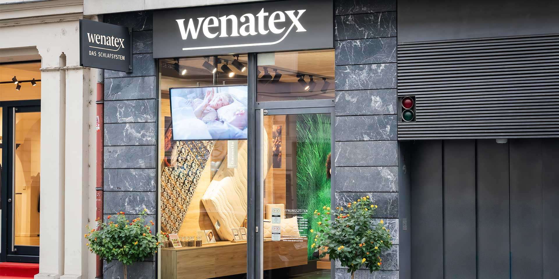 Außenansicht eines Geschäfts mit Schaufenster, Pflanzen und einem großen Bildschirm.