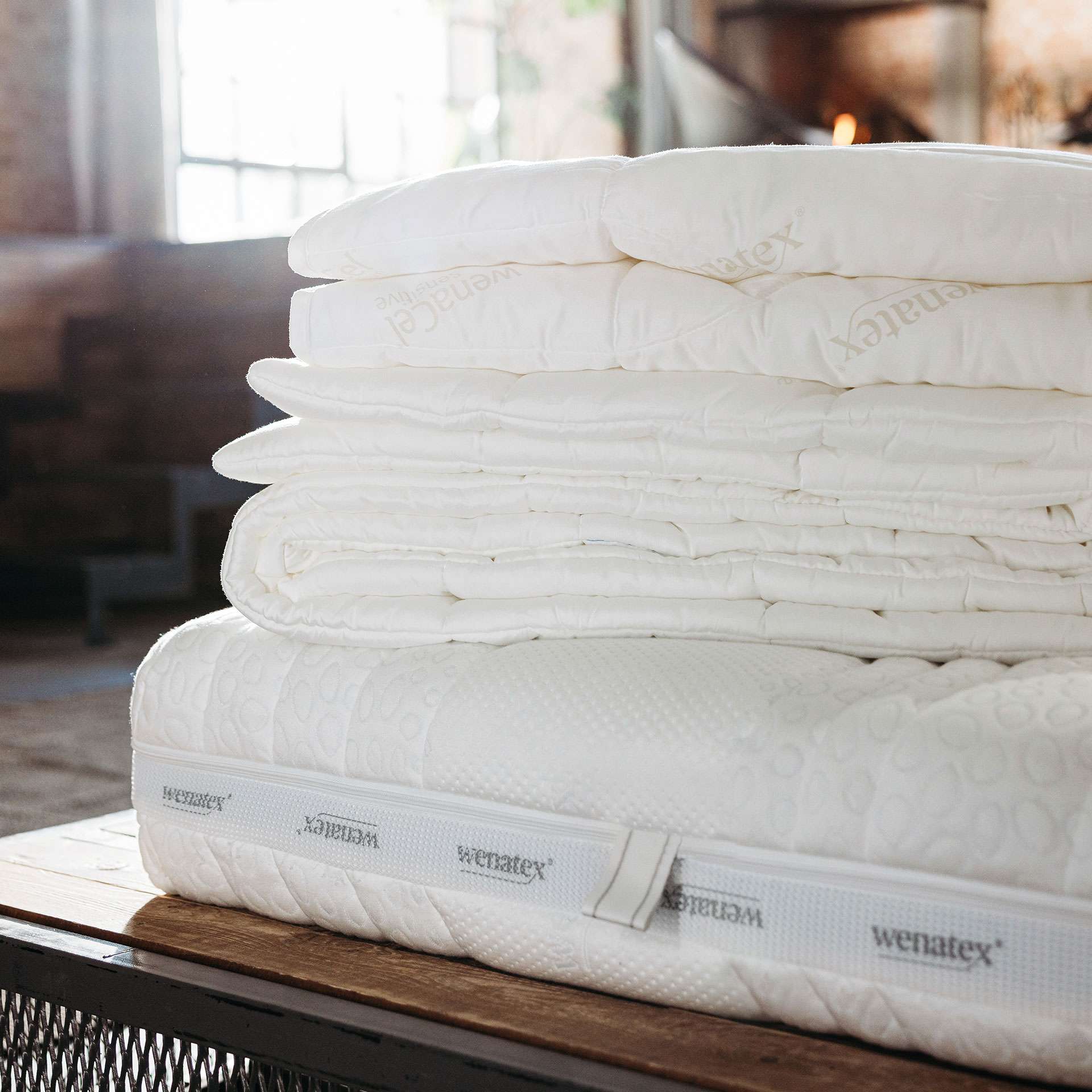 Stack of Wenatex mattress toppers and blankets on a wooden table in a cozy room.