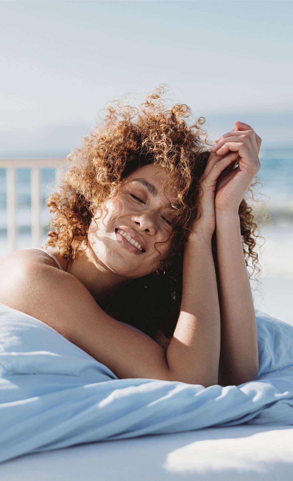 Frau mit lockigem Haar entspannt sich lächelnd auf einem Bett mit blauer Bettdecke am Meer.