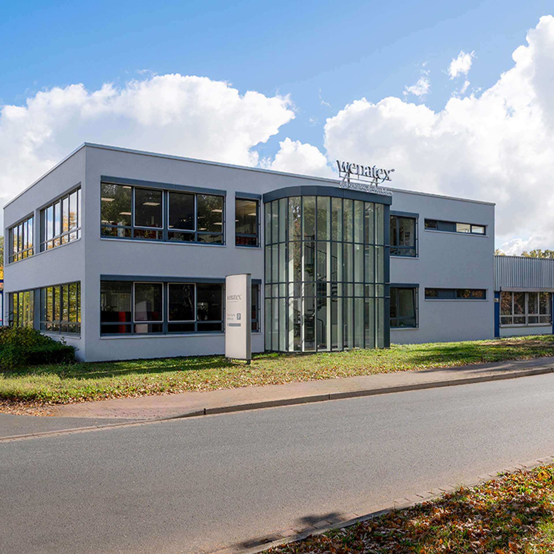 Modernes Bürogebäude mit Glasfront, umgeben von Bäumen und einer Straße im Vordergrund. Wolken am Himmel.