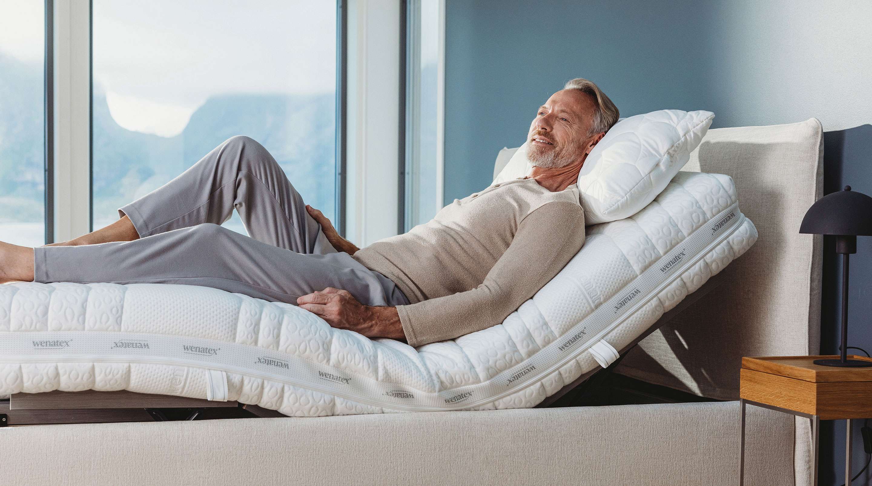 An older man relaxes on an adjustable bed with a headboard and mattress in a bright room.