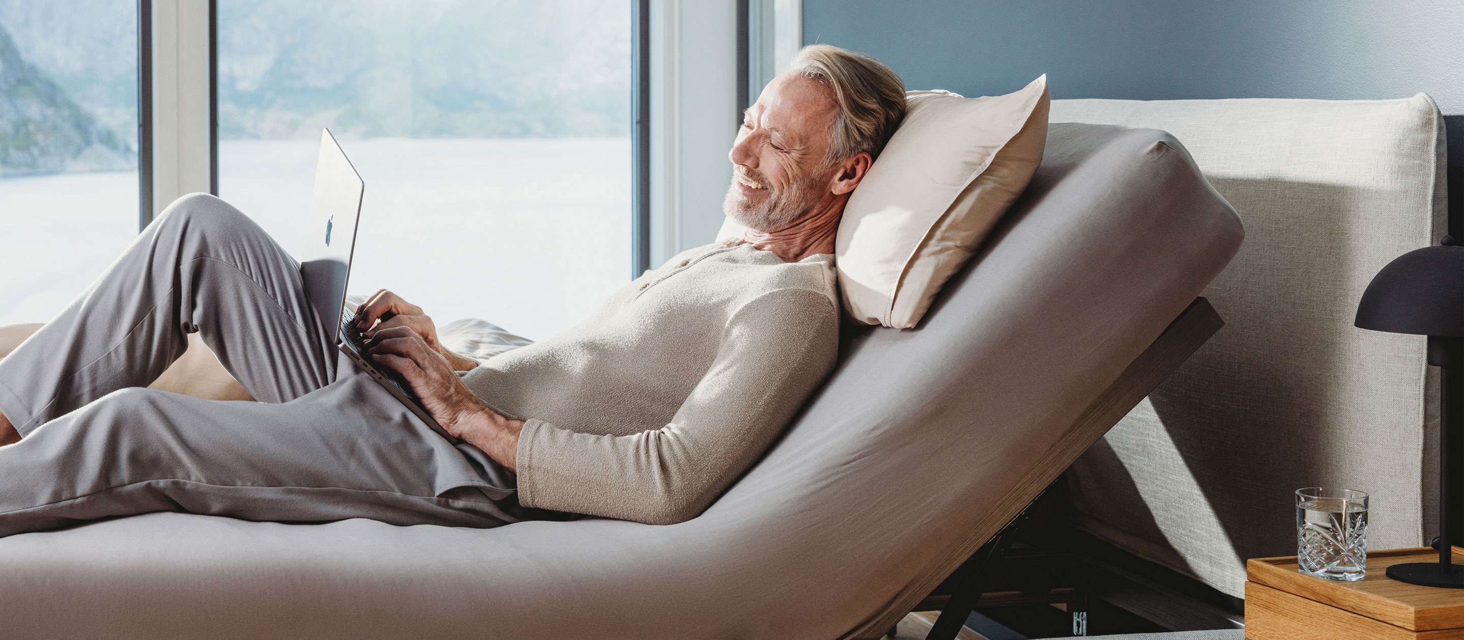 Man relaxes on a bed with a laptop, smiling and enjoying the view through a large window.