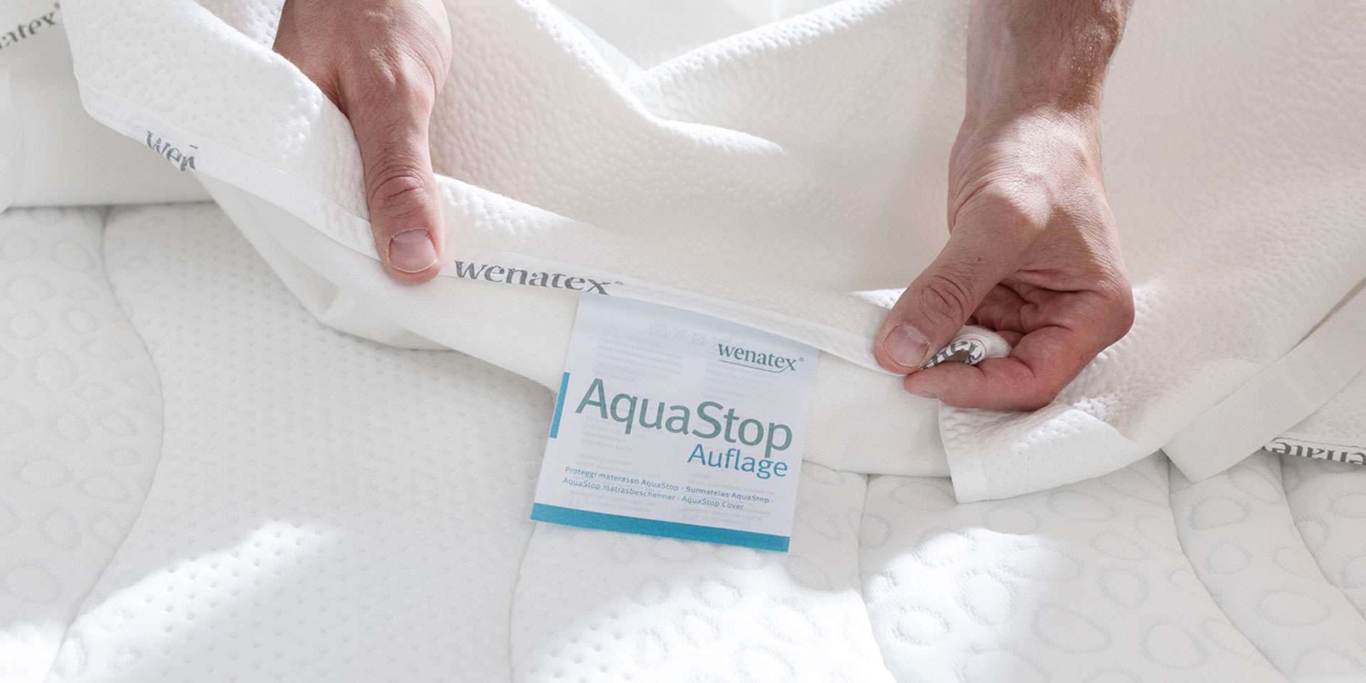 Hände halten eine Wenatex AquaStop Auflage, die auf einer Matratze platziert ist.