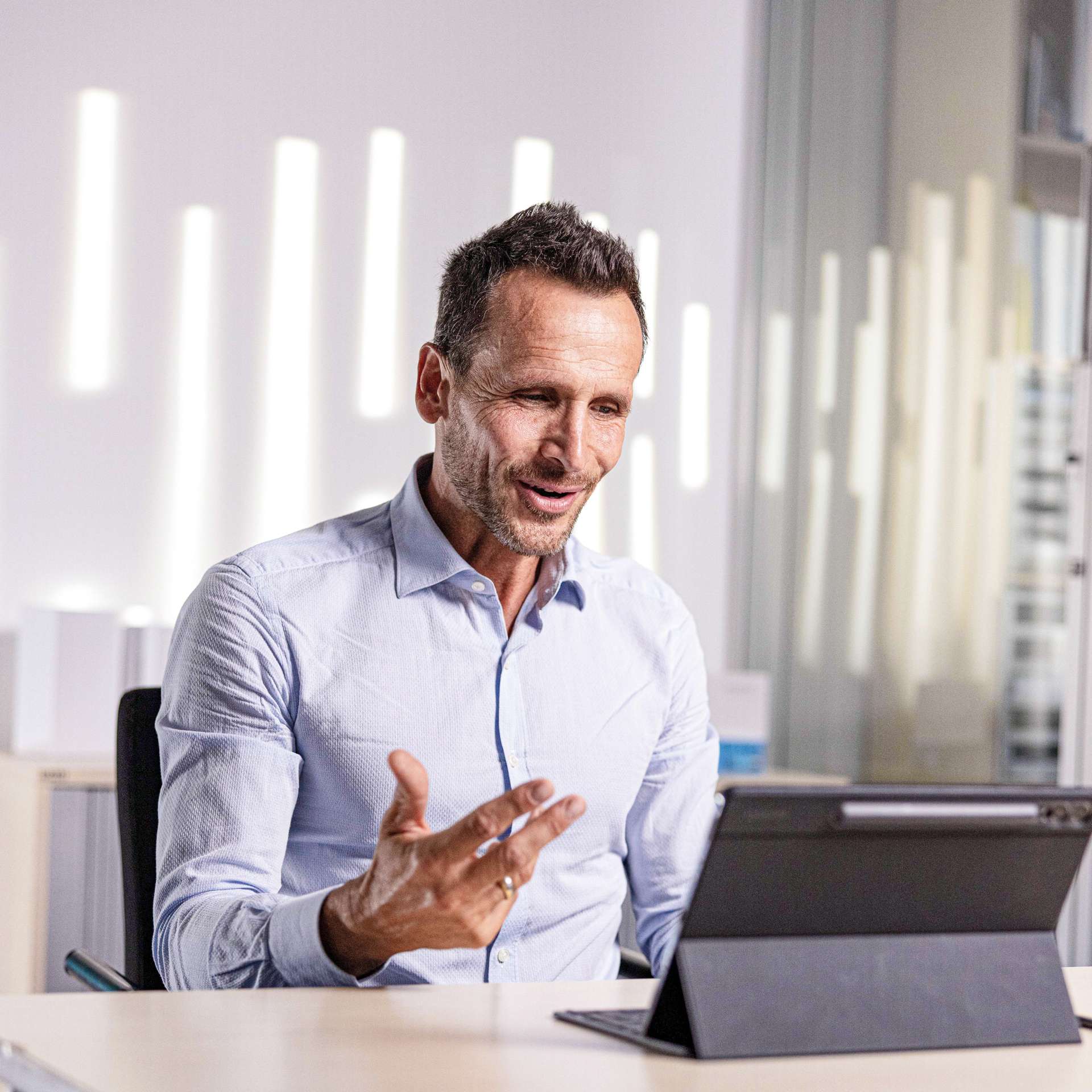 Man in a shirt smiling while speaking into a tablet during an online consultation. Bright, modern office setting.