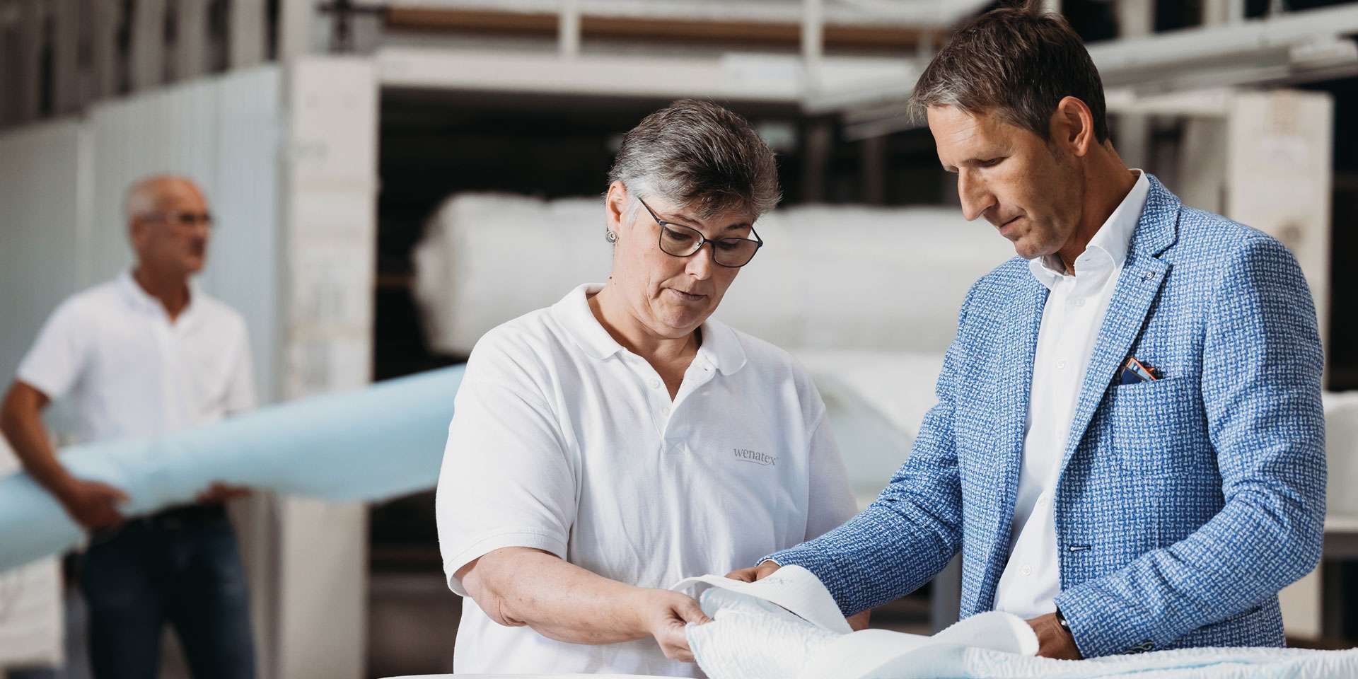 Zwei Personen prüfen Materialien in einer Produktionsumgebung, während eine weitere Person im Hintergrund arbeitet.