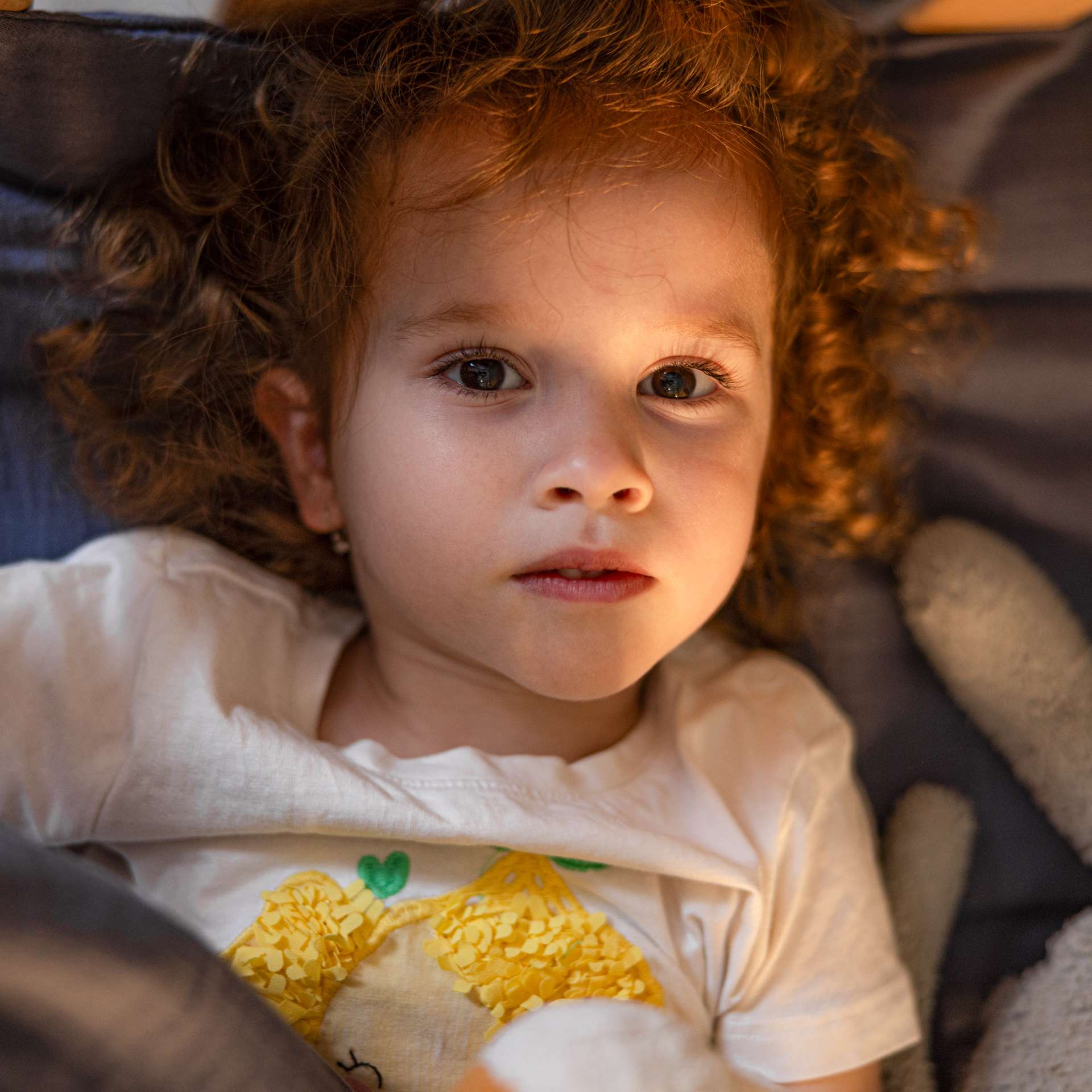 A small child with curly hair lies on a bed, looking directly at the camera.