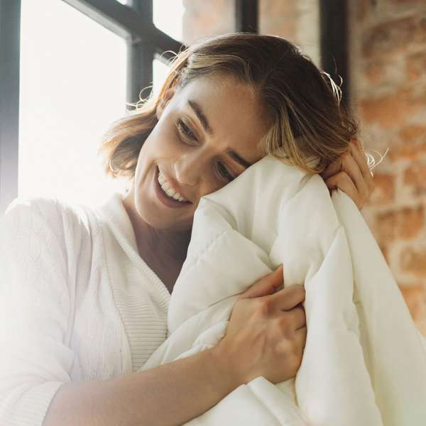 Woman hugs a light duvet and smiles happily in a bright room.