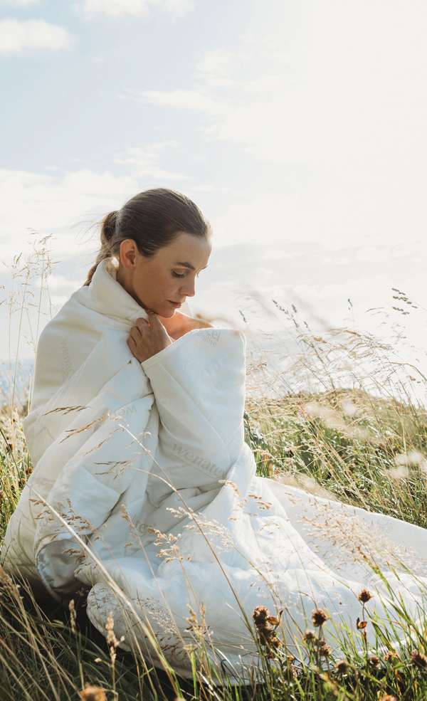 Woman wraps herself in a light blanket, sitting in a blooming meadow under a blue sky.