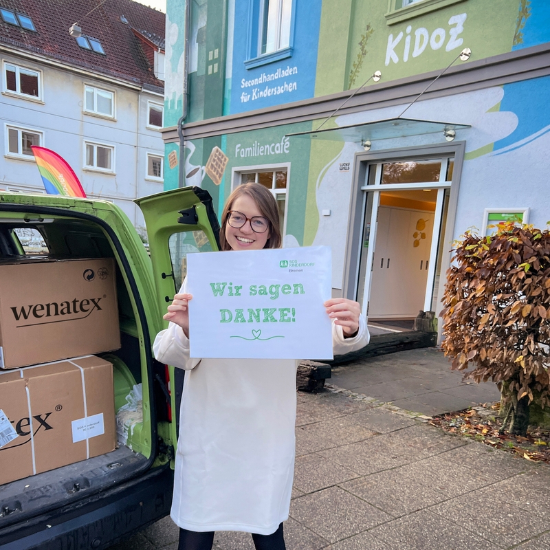 Frau hält ein Schild mit der Aufschrift "Wir sagen DANKE!" vor einem Gebäude mit buntem Wanddesign.
