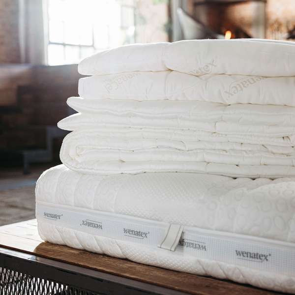 Stapelt von Wenatex Matratzenauflagen und Bettdecken auf einem Holztisch in einem gemütlichen Raum.