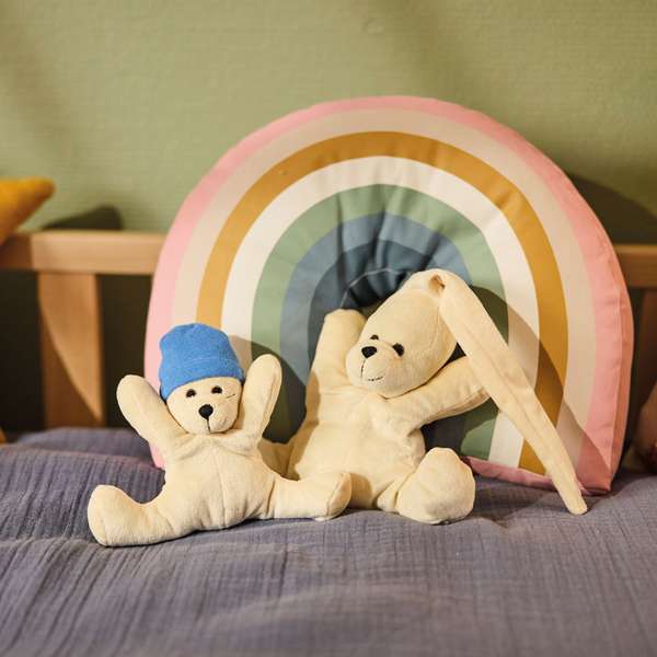 Zwei Stoffbären auf einem Bett mit einem bunten Regenbogen-Kissen im Hintergrund.