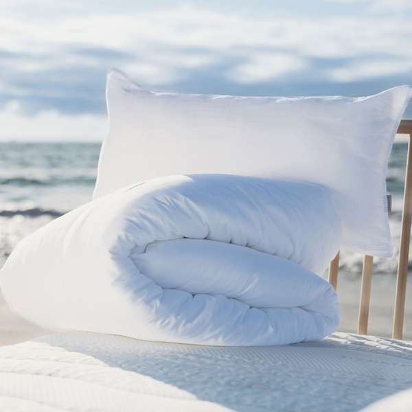 Wenatex Kissen und Bettdecke auf einem Bett am Strand mit Meer im Hintergrund.